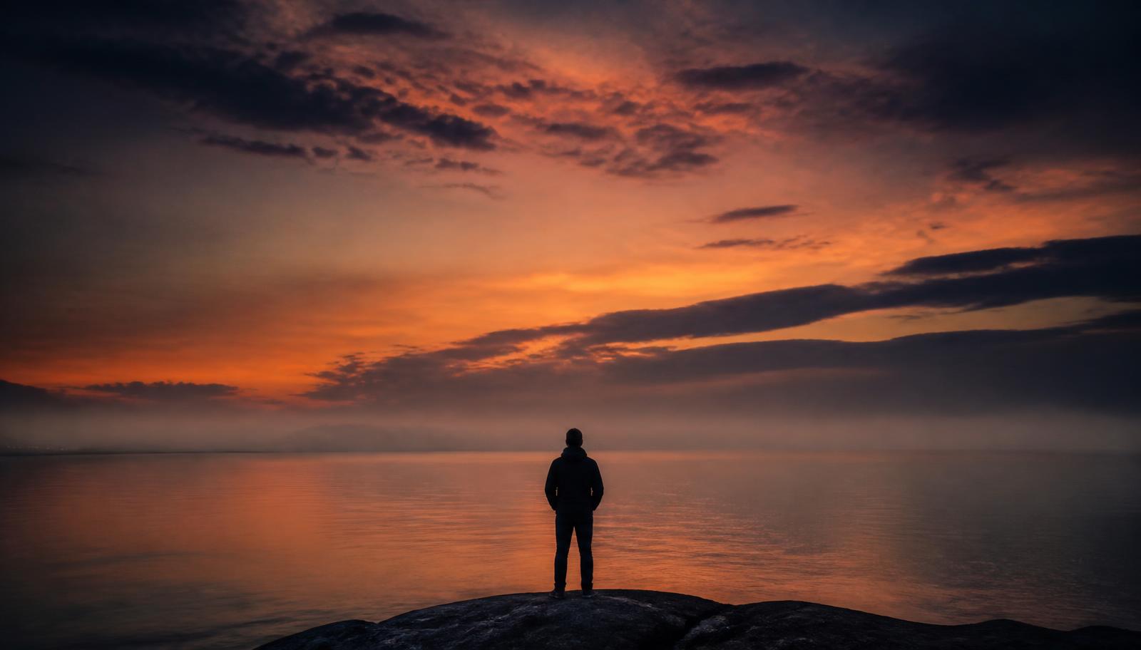 Silueta de una persona frente a un amanecer inmenso sobre el mar en calma, tonos índigo y ámbar